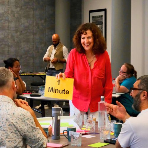 Jane stands in a room of people seated at tables in a workshop style. She holds a sign that says 1 minute.